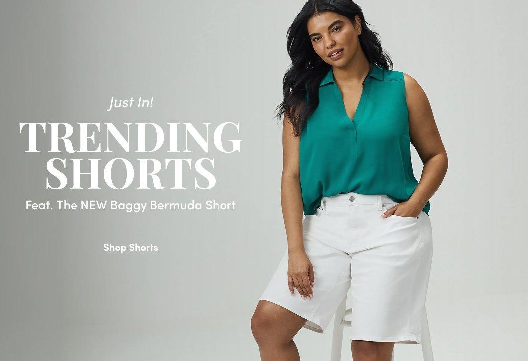 A promotional banner with a white background. A woman with dark hair pulled back stands leaning against a white surface with one hand resting on it. She wears a sleeveless teal V-neck blouse tucked into white Bermuda shorts, with white sneakers. White and dark text overlaid on the left reads 'just in!' in small italic letters, followed by 'TRENDING SHORTS' in large bold text, then 'Feat. The NEW Baggy Bermuda Short' in smaller text. Below that is a dark rectangular button with white text reading 'Shop Shorts'.