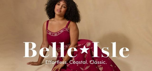 A wide promotional banner with a warm sandy beige background featuring two women with voluminous curly hair posed on either side. On the left, a woman stands with one hand raised behind her head, wearing a short-sleeve V-neck midi dress in cream with a pink and magenta floral print and ruffle sleeves. On the right, a woman sits and leans to one side, wearing a deep red sleeveless crop top with white trim detail paired with a matching red printed maxi skirt. Centered between them in large white serif text reads 'Belle*Isle' with a star in place of the space, and below it in smaller white text, 'Effortless. Coastal. Classic.'