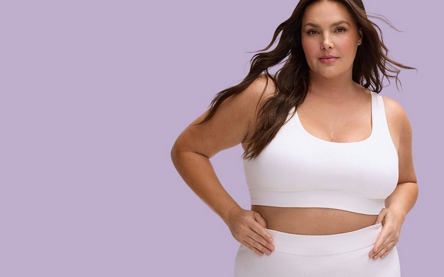 A vertical promotional banner for the Seamless Rib Collection featuring a model against a light lavender background. She is wearing a white ribbed seamless scoop-neck bra and matching high-waisted ribbed bottoms, with her hands resting on her hips.