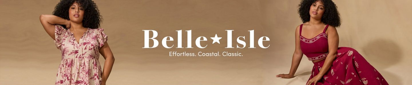 A wide promotional banner with a warm sandy beige background featuring two women with voluminous curly hair posed on either side. On the left, a woman stands with one hand raised behind her head, wearing a short-sleeve V-neck midi dress in cream with a pink and magenta floral print and ruffle sleeves. On the right, a woman sits and leans to one side, wearing a deep red sleeveless crop top with white trim detail paired with a matching red printed maxi skirt. Centered between them in large white serif text reads 'Belle*Isle' with a star in place of the space, and below it in smaller white text, 'Effortless. Coastal. Classic.'