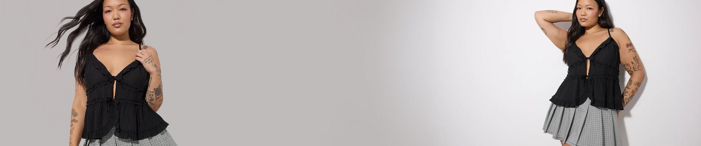 'A wide promotional banner featuring the same plus-size woman with long straight auburn hair shown from two angles against a weathered cream and beige textured wall with a graveled ground. She wears smoky eye makeup, small drop earrings, a fitted black square-neck tank top with a decorative black floral applique detail on one strap, and olive green leggings with an all-over black botanical and peacock jacquard print. On the left she faces slightly forward, and on the right she is seated and angled toward the camera.'