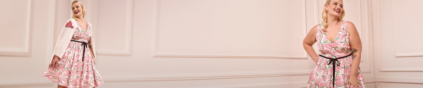 'A wide promotional banner featuring the same plus-size woman with short wavy blonde hair and red lipstick shown from two angles against a soft blush pink paneled wall interior. She wears small gold earrings and a white fit-and-flare midi dress printed with large pink watermelon slice illustrations with green rinds and black seeds, cinched at the waist with a black ribbon tie. On the left she wears the dress paired with a white short-sleeve cropped jacket featuring a large watermelon applique on the back, laughing and looking to the side. On the right she stands with one hand on her hip smiling upward, with a tattoo visible on her right forearm.'