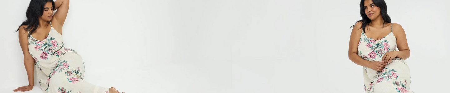 'A wide promotional banner featuring the same plus-size woman with long straight auburn hair shown from two angles against a weathered cream and beige textured wall with a graveled ground. She wears smoky eye makeup, small drop earrings, a fitted black square-neck tank top with a decorative black floral applique detail on one strap, and olive green leggings with an all-over black botanical and peacock jacquard print. On the left she faces slightly forward, and on the right she is seated and angled toward the camera.'