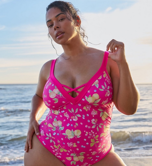 A woman with dark hair pulled back in a damp updo stands at the beach with calm ocean water and a partly cloudy sky visible behind her. She looks downward with one hand raised near her shoulder. She wears a bright pink one-piece swimsuit with an allover fruit print featuring lemons, blueberries, strawberries, cherries, and green leaves, a deep V-neckline with crisscross strap detail at the center front, and wide shoulder straps. She wears a red floral earring visible on her left ear.