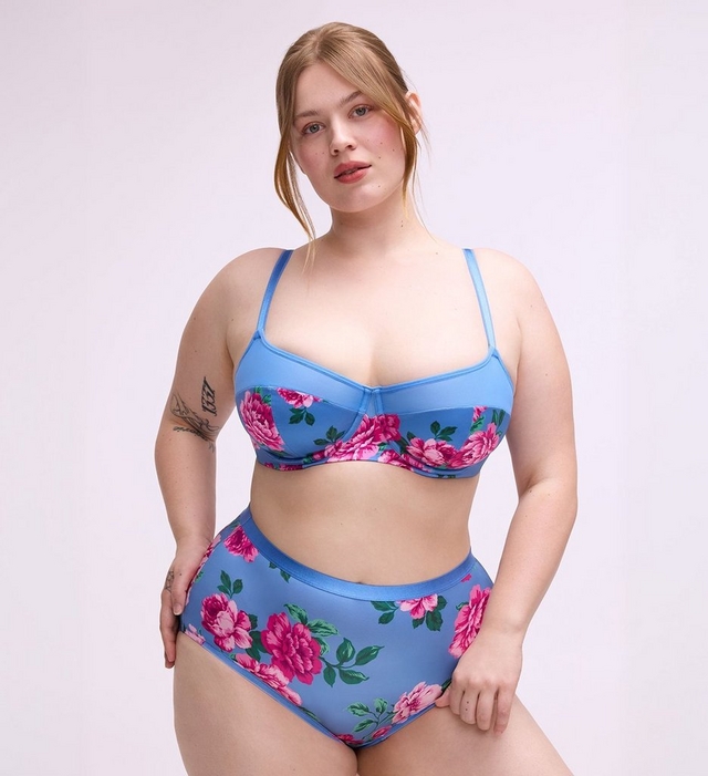 A woman with straight reddish-blonde hair pulled back loosely stands against a plain light gray background, looking directly at the camera.A woman with straight reddish-blonde hair pulled back loosely stands against a plain light gray background, looking directly at the camera. She wears a two-piece bikini set in blue with a large pink peony and green leaf print. The top is an underwire balconette style with thin straps, and the bottoms are high-waist briefs with a solid blue waistband. She has tattoos on her left upper arm and right hand, and one hand rests at her hip.