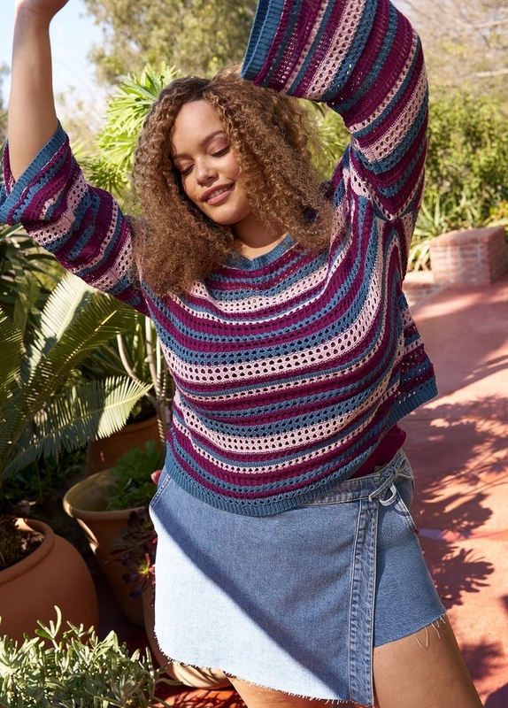 A woman with long curly reddish-brown hair stands outdoors in a sunny garden setting with tropical plants, terracotta pots, and a brick ledge visible in the background. She has both arms raised above her head, eyes closed, and is smiling. She wears an open-knit striped sweater in horizontal bands of teal, magenta, and blush pink, paired with a light blue denim mini skirt with a raw frayed hem.