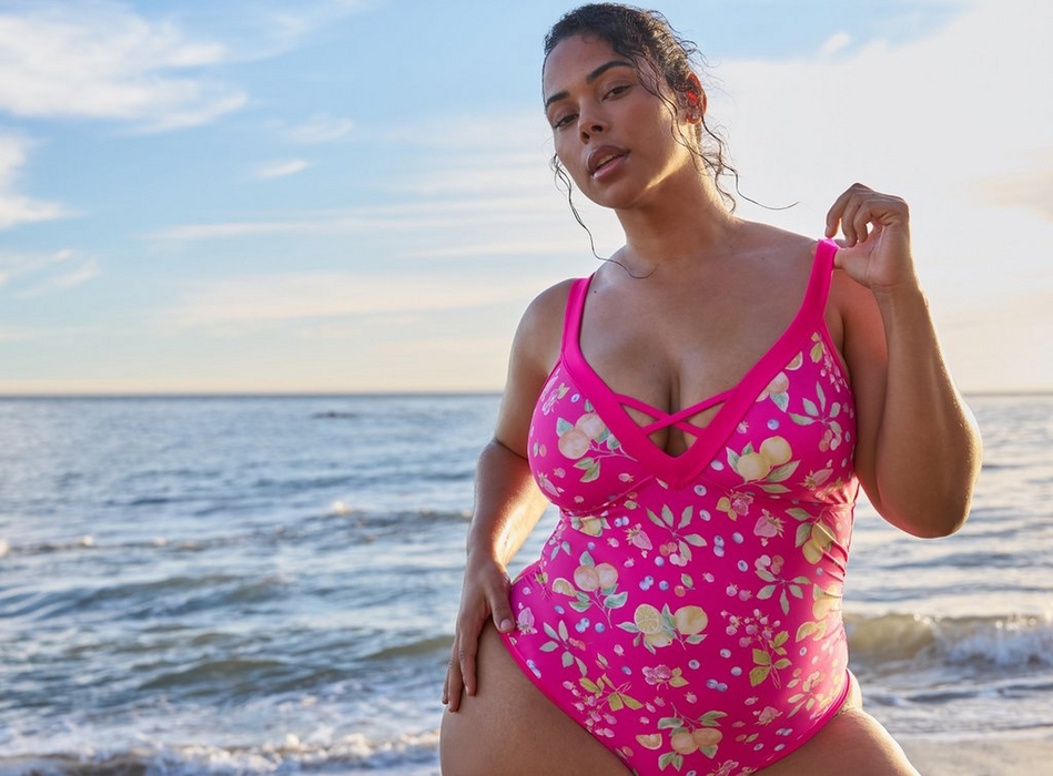 A woman with dark hair pulled back in a damp updo stands at the beach with calm ocean water and a partly cloudy sky visible behind her. She looks downward with one hand raised near her shoulder. She wears a bright pink one-piece swimsuit with an allover fruit print featuring lemons, blueberries, strawberries, cherries, and green leaves, a deep V-neckline with crisscross strap detail at the center front, and wide shoulder straps. She wears a red floral earring visible on her left ear.