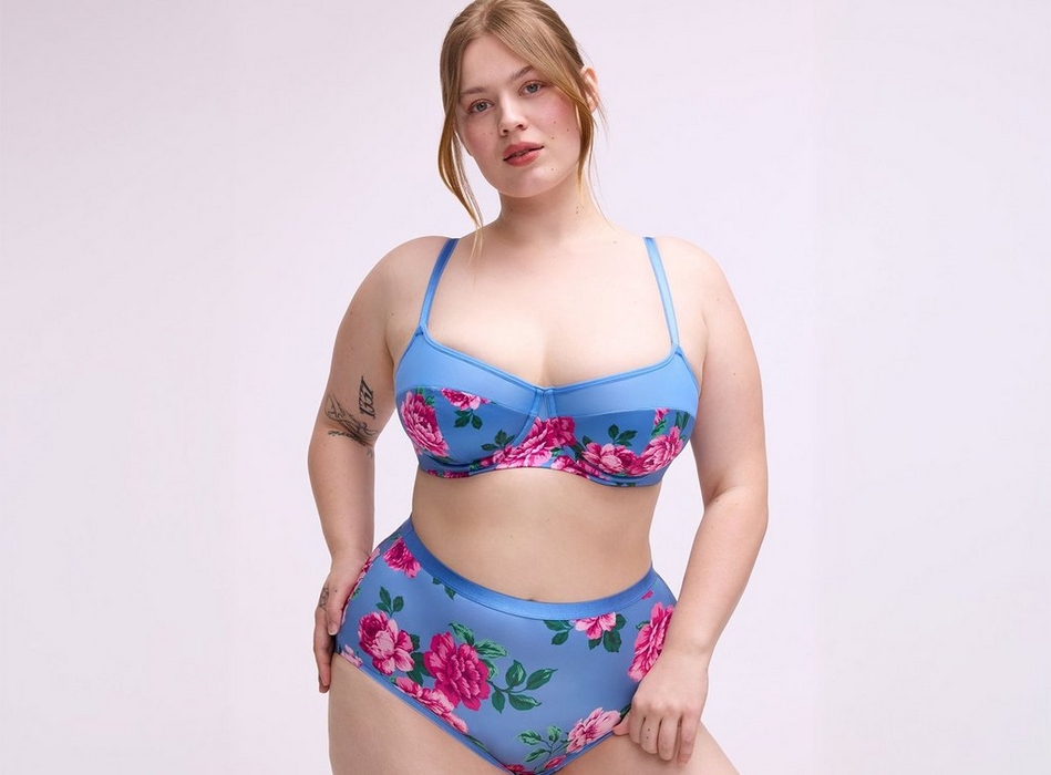 A woman with straight reddish-blonde hair pulled back loosely stands against a plain light gray background, looking directly at the camera.A woman with straight reddish-blonde hair pulled back loosely stands against a plain light gray background, looking directly at the camera. She wears a two-piece bikini set in blue with a large pink peony and green leaf print. The top is an underwire balconette style with thin straps, and the bottoms are high-waist briefs with a solid blue waistband. She has tattoos on her left upper arm and right hand, and one hand rests at her hip..