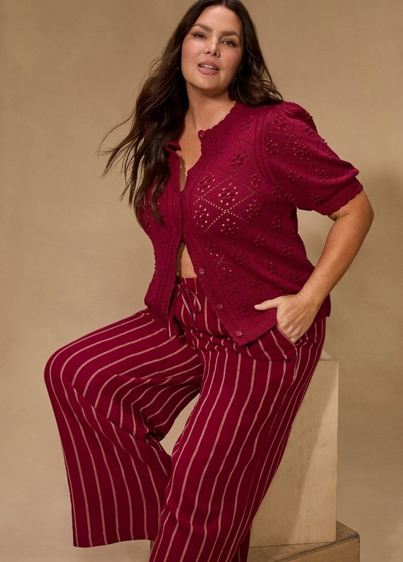 A woman with long dark wavy hair sits on the edge of a white cube prop with one foot raised, hand in pocket, against a warm tan beige background. She wears a deep magenta short-sleeve button-front cardigan with an open-knit pointelle and bobble texture and scalloped neckline, paired with wide-leg pants in deep magenta with thin cream vertical stripes and a tie waist.