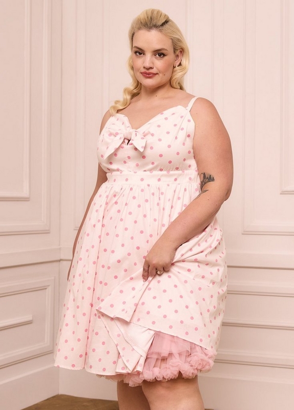  A woman with blonde hair styled in a vintage-inspired updo with a side wave stands against a soft blush pink paneled wall. She wears a sleeveless fit-and-flare mini dress in white with large pink polka dots, a sweetheart neckline with a bow detail at the center front, and a full skirt with a pink tulle ruffle petticoat peeking out at the hem. She has a tattoo on her right upper arm and wears a ring on her right hand. A small earring is visible.