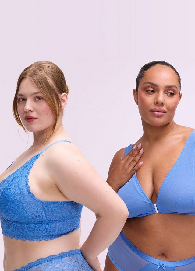 A woman with straight reddish-blonde hair pulled back loosely stands against a plain light gray background, looking directly at the camera.A woman with straight reddish-blonde hair pulled back loosely stands against a plain light gray background, looking directly at the camera. She wears a two-piece bikini set in blue with a large pink peony and green leaf print. The top is an underwire balconette style with thin straps, and the bottoms are high-waist briefs with a solid blue waistband. She has tattoos on her left upper arm and right hand, and one hand rests at her hip.