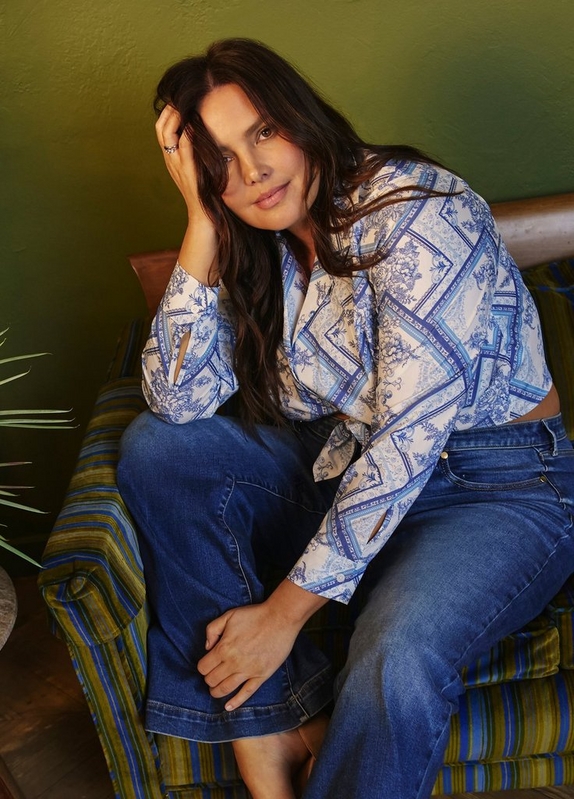 'A woman with long dark wavy hair sits on a green and blue striped cushioned chair or ottoman, leaning her head on one hand and looking at the camera with a slight smile. She wears a white and blue scarf-print blouse with long sleeves, a V-neckline with a keyhole detail, and bell cuffs, paired with medium-wash blue wide-leg jeans. She wears rings on her right hand and tan sandals. A green-painted wall and partial palm leaf are visible in the background.