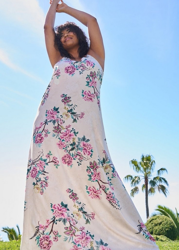 'A low-angle shot looking up at a woman with short dark curly hair standing outdoors with both arms raised above her head, eyes closed and face tilted upward. She wears a white sleeveless maxi dress with an allover large-scale pink floral and branch print with teal and green leaves. A bright blue sky, palm tree, and green hedges are visible in the background.