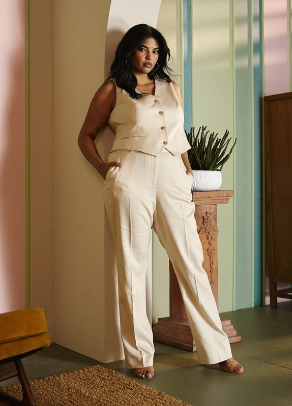 A woman with long dark wavy hair and gold hoop earrings stands leaning against a pale pillar in a stylized interior space with pink and green paneled walls, an olive green floor, a woven rug, a mustard yellow chair, and a terracotta pedestal with a cactus in a white pot. She looks off to one side with one hand in her pocket. She wears a cream sleeveless button-front vest with gold buttons and a V-neckline with lapels, paired with matching cream wide-leg trousers with front pleats, and tan strappy heeled sandals.