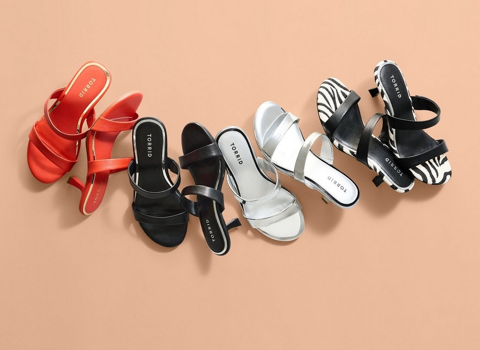 A flat lay of four strappy heeled sandals arranged in a row against a peach-toned background with soft shadow detail. From left to right: a cream sandal with thin crisscross straps and gold hardware, a red sandal with thin crisscross straps and gold hardware, another red sandal with thin crisscross straps and gold hardware shown from a slightly different angle, and a black sandal with thin crisscross straps and silver hardware. All sandals have square toes, slender stiletto heels, and tan insoles with 'TORRID' printed on each.