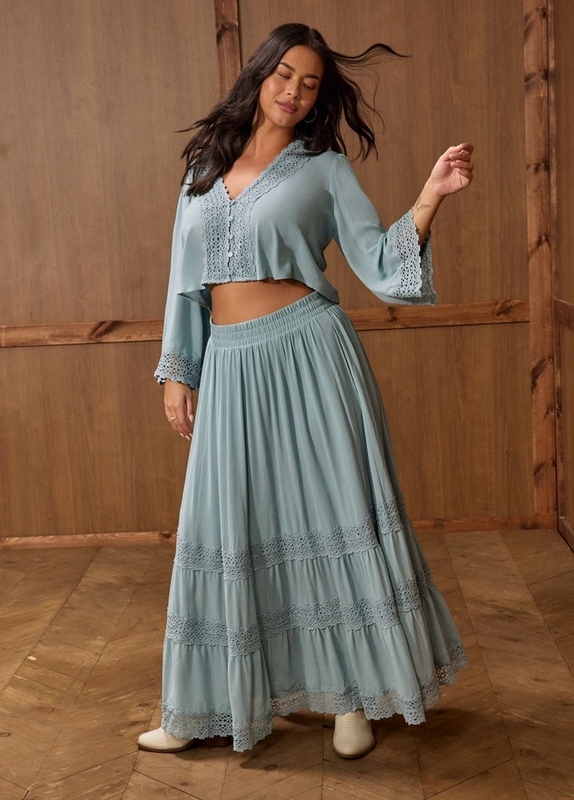 A woman with long dark hair poses indoors wearing a dusty blue boho two-piece set consisting of a cropped long-sleeve top with a V-neckline, button front, and crochet lace trim at the collar and cuffs, paired with a matching full-length tiered maxi skirt featuring multiple rows of crochet lace detailing along the hem. She wears cream ankle boots and gold hoop earrings and has a small tattoo on her wrist. The background is a warm brown wood-paneled wall, and she stands on a herringbone hardwood floor.