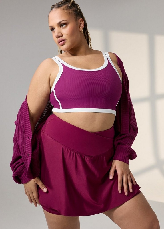 A woman with long braided hair pulled up poses against a light gray background with soft window light. She wears a magenta ribbed sports bra with white contrast trim along the neckline, straps, and hem, paired with a matching magenta athletic skort with a crossover waistband. A ribbed magenta knit cardigan is draped off her shoulders. She wears small gold earrings.