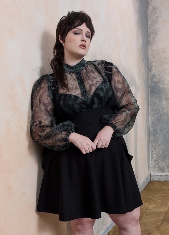 A woman with short dark hair and wispy bangs leans against a textured plaster wall in muted peach and gray tones. She wears a dark green sheer floral organza blouse with voluminous long sleeves, a high neckline with small buttons, and black hardware buckle details at the shoulders, layered over a black sleeveless pinafore dress with a fitted bodice and a flared skirt. She wears small drop earrings and multiple rings and stands on a herringbone wood floor, looking downward with a moody expression.