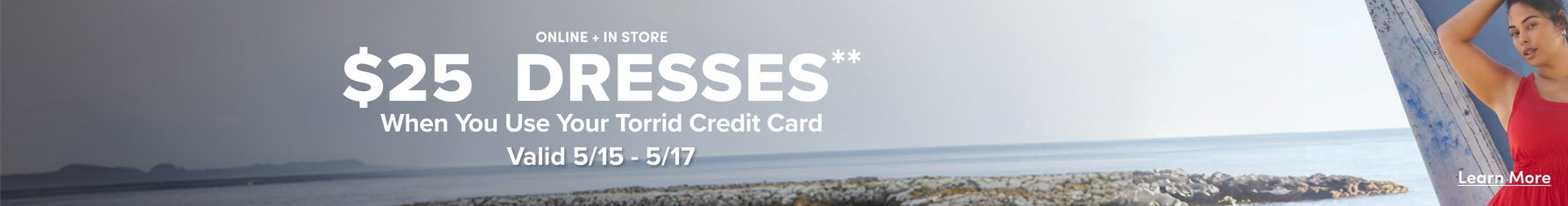 A wide horizontal promotional banner with a muted coastal landscape background showing a rocky shoreline, calm gray-blue ocean, and distant mountains under an overcast sky. White text in the center reads 'ONLINE + IN STORE' in small caps above large bold text reading '$25 DRESSES**', followed by 'When You Use Your Torrid Credit Card' and 'Valid 5/15 - 5/17' in smaller bold text. On the far right, a woman wearing a red sleeveless dress is partially visible, leaning against a weathered wooden post. An underlined 'Learn More' link appears in the lower right corner.