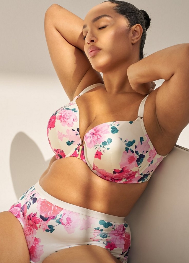 A close-up vertical image of a woman with dark hair pulled back in a bun, reclining against a light beige surface with a soft gray background and faint shadow. Her eyes are closed and both arms are raised behind her head. She wears a matching floral lingerie set with a cream base printed with pink, red, and teal roses and floral motifs. The underwire bra features a gold front clasp and adjustable cream straps, and the matching high-waist briefs have a plain cream waistband. Soft natural light falls across her from one side.