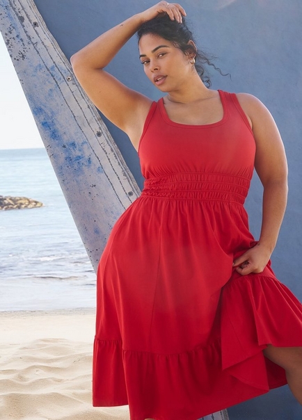 A woman with dark hair loosely pulled back poses outdoors at a beach, leaning against a weathered wooden post in front of a blue-gray wall with the ocean and sandy shoreline visible in the background. She wears a red sleeveless midi dress with a square neckline, wide shoulder straps, a smocked waistband, and a tiered ruffle hem. She has one hand raised to her head and the other resting on her hip. She wears small gold hoop earrings and rings on her fingers.