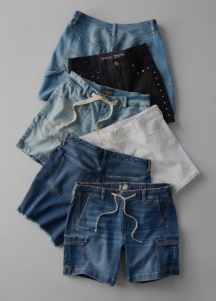 A flat-lay product image on a light gray background showing six pairs of denim and casual shorts fanned and stacked in an overlapping arrangement. From top to bottom they include a light wash blue denim mini short, a black denim short with silver studded detailing along the side and a visible Torrid label, a light blue denim short with a drawstring waist, a white denim short with a button fly, a dark indigo denim short with raw-cut frayed hem, and a medium wash blue denim cargo short with a drawstring waist and cargo pockets.