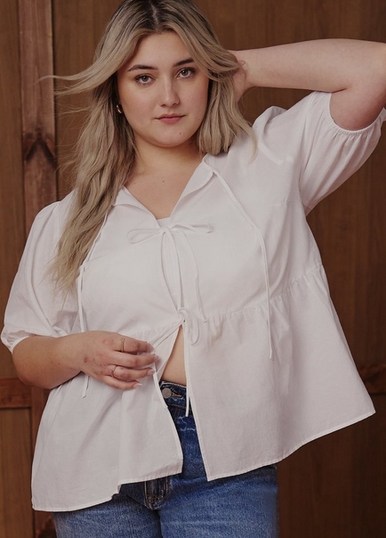 A woman with long wavy blonde hair poses in front of a warm brown wood-paneled background, with one hand raised behind her head. She wears a white short-sleeve oversized top with a collared V-neckline, a front tie closure with two bows, a peplum ruffle hem, and elasticized sleeve openings, worn open over a white bralette. The top is partially open at the front hem. She wears blue jeans underneath and has small gold earrings and two rings on her fingers.