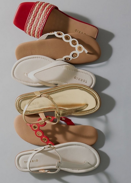 A flat-lay product image on a light gray background showing five sandals arranged in a vertical overlapping stack, all branded with the Torrid name on the insole. From top to bottom they include a red square-toe slide with woven raffia and red rope braid across the toe strap, a white T-strap sandal with gold grommet chain detail and a tan footbed, a simple white thong flip flop with a smooth strap and white footbed, a gold metallic T-strap sandal with a gold footbed, and a red T-strap sandal with gold grommet chain detail and a tan footbed, and a white T-strap sandal with a gold buckle detail and cream footbed.