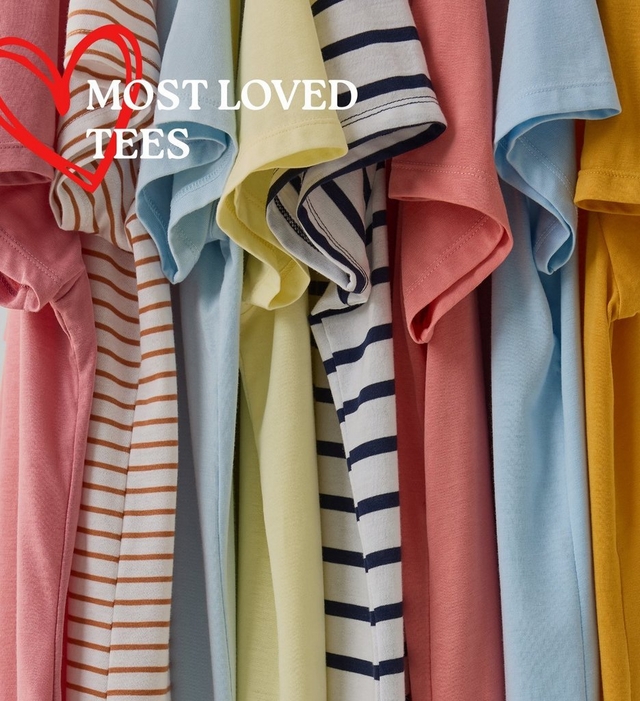 A close-up image of multiple t-shirts hanging tightly together, showing a colorful range of sleeve styles and fabrics. Visible colors and patterns from left to right include pink, white with brown horizontal stripes, light blue, pale yellow, white with navy blue stripes, coral pink, light blue, and mustard yellow. In the upper left corner, a red hand-drawn heart outline is overlaid with bold white text reading 'MOST LOVED TEES'.
