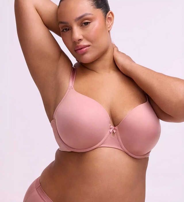 A woman with dark hair pulled back poses against a soft lavender-gray background, looking directly at the camera with one arm raised above her head and the other hand resting at her neck. She wears a smooth mauve-pink T-shirt style underwire bra with a small bow detail at the center front gore and thin matching straps.