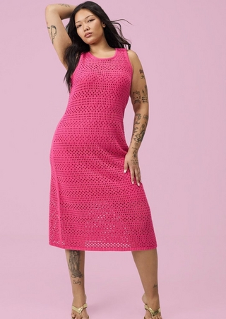 A woman with long dark hair poses against a solid pink background, one arm raised behind her head and the other resting at her hip. She wears a hot pink sleeveless midi dress in an open-weave crochet knit with a round neckline and a slightly A-line silhouette. She wears gold strappy heeled sandals. Both of her arms and legs are heavily tattooed with a variety of designs including florals, figures, and text.