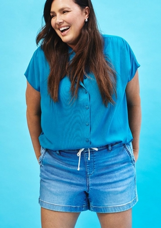 A woman with long dark hair laughs openly against a bright turquoise blue background. She wears a teal short-sleeve button-front blouse with a collared neckline and cap sleeves, paired with light wash denim shorts with a white drawstring waist. She wears small crystal stud earrings and has both hands in her pockets.