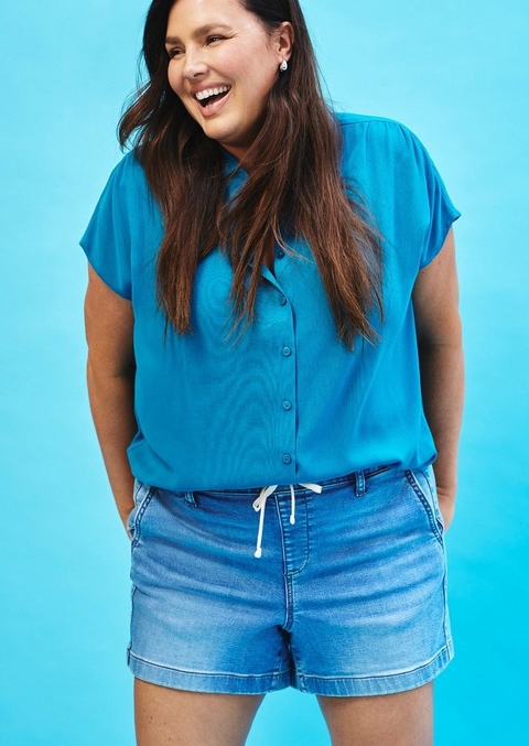 A woman with long dark hair laughs openly against a bright turquoise blue background. She wears a teal short-sleeve button-front blouse with a collared neckline and cap sleeves, paired with light wash denim shorts with a white drawstring waist. She wears small crystal stud earrings and has both hands in her pockets.