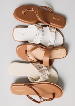 A flat-lay product image on a light gray background showing four sandals arranged in a vertical overlapping stack, all featuring warm neutral tones and Torrid branding on the insoles. From top to bottom they include a tan thong flip flop with a braided leather T-strap and gold hardware detail, a white double-strap slide with contrast topstitching and a gold square buckle on a tan sole, a tan strappy T-strap sandal with a crisscross toe strap and a blush footbed, and a cream woven raffia slide with a clear buckle detail and a cream canvas footbed, with a second tan braided thong flip flop partially visible at the bottom.