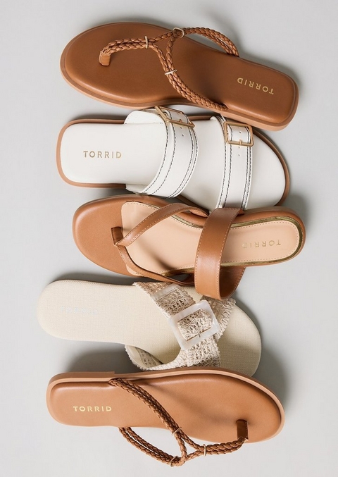 A flat-lay product image on a light gray background showing four sandals arranged in a vertical overlapping stack, all featuring warm neutral tones and Torrid branding on the insoles. From top to bottom they include a tan thong flip flop with a braided leather T-strap and gold hardware detail, a white double-strap slide with contrast topstitching and a gold square buckle on a tan sole, a tan strappy T-strap sandal with a crisscross toe strap and a blush footbed, and a cream woven raffia slide with a clear buckle detail and a cream canvas footbed, with a second tan braided thong flip flop partially visible at the bottom.