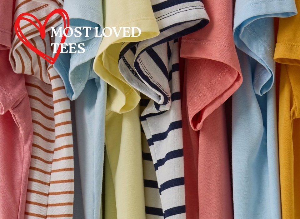 A close-up image of multiple t-shirts hanging tightly together, showing a colorful range of sleeve styles and fabrics. Visible colors and patterns from left to right include pink, white with brown horizontal stripes, light blue, pale yellow, white with navy blue stripes, coral pink, light blue, and mustard yellow. In the upper left corner, a red hand-drawn heart outline is overlaid with bold white text reading 'MOST LOVED TEES'.