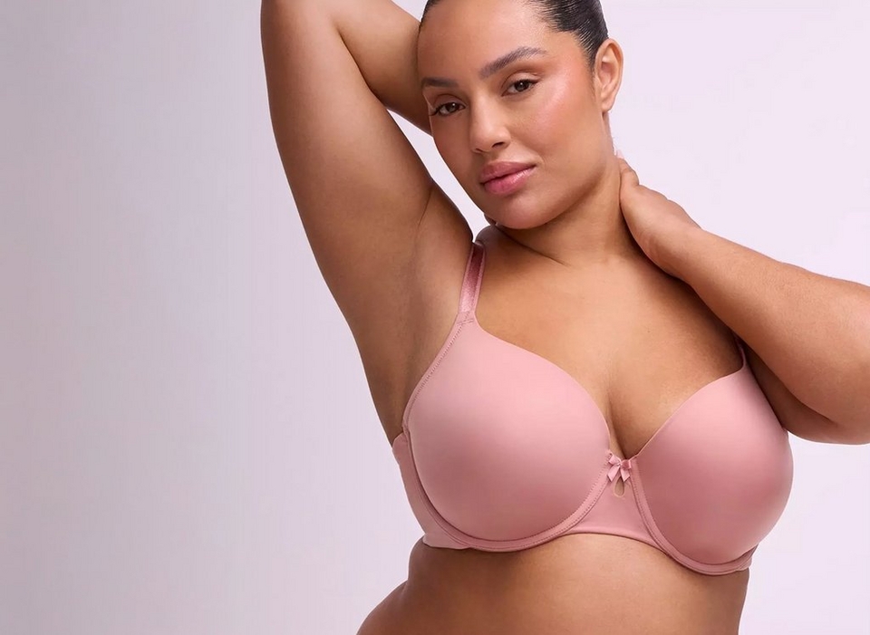 A woman with dark hair pulled back poses against a soft lavender-gray background, looking directly at the camera with one arm raised above her head and the other hand resting at her neck. She wears a smooth mauve-pink T-shirt style underwire bra with a small bow detail at the center front gore and thin matching straps.