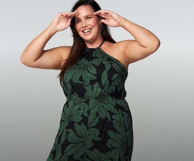 A woman with long straight dark hair smiles broadly at the camera with both hands raised near her temples against a light gray background. She wears a black sleeveless halter-neck jumpsuit with a bold large-scale green tropical floral print, featuring a gathered halter neckline, an elasticized waist, and wide-leg pants. She wears small hoop earrings and a small wrist tattoo is visible on one arm.