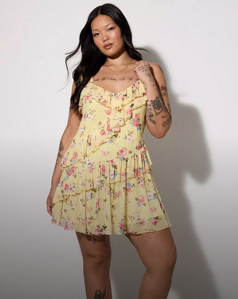 A woman with long straight dark hair poses against a plain white background, one hand raised to her chest. She wears a pale yellow spaghetti-strap mini dress printed with pink roses, blue flowers, and green leaves, featuring a ruffled sweetheart neckline, a cascading ruffle panel down the center front, and two tiered ruffle layers on the skirt. She has extensive tattoos visible on both arms, her chest, and her legs. She wears a delicate gold chain necklace.