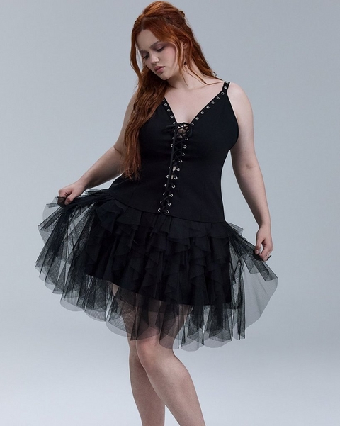 A woman with long wavy red hair looks downward against a soft gray background, both hands gently holding out the skirt of her dress. She wears a black sleeveless mini dress with a deep V-neckline featuring a corset-style lace-up front with silver grommet hardware, thin straps with grommet detailing at the shoulders, and a voluminous layered tulle skirt with a ruffled hem. She wears rings on both hands and small earrings.