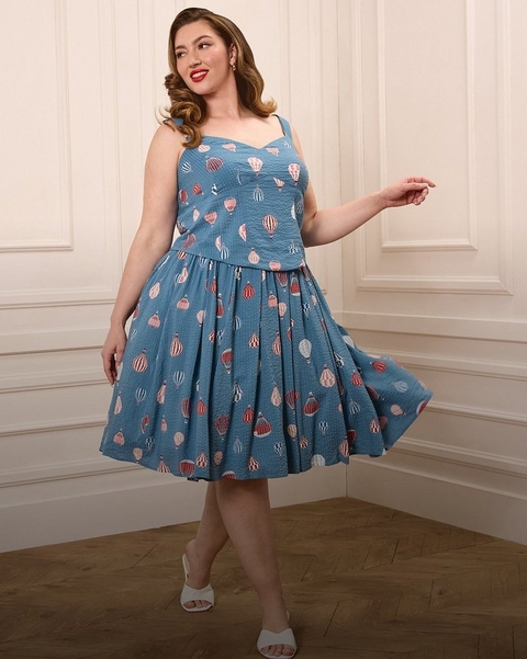 A woman with vintage-style side-swept waves and bold red lips smiles and poses in a bright white paneled room with herringbone hardwood floors. She wears a matching two-piece seersucker set in medium blue printed with colorful hot air balloons in red, white, and pink, consisting of a fitted sleeveless crop top with a sweetheart neckline and wide straps, paired with a full pleated midi skirt that flares as she twirls. She wears white open-toe mule heels and small pearl drop earrings.