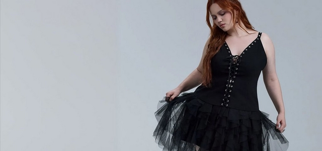 A model with long auburn hair against a light grey background wearing a black lace-up corset-style mini dress with grommet hardware and a layered black tulle ruffled skirt, holding the skirt out to the side.