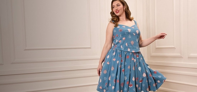 A model with pin-up style wavy brown hair and red lipstick poses against a white paneled wall wearing a blue fit-and-flare dress with a hot air balloon print, sweetheart neckline, wide straps, and a full pleated skirt.