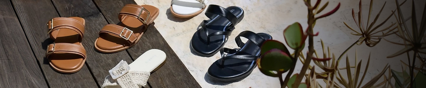 A wide promotional banner featuring five pairs of summer sandals arranged in a flat lay on a wooden deck and stone surface with tropical succulent foliage in the lower right corner. Styles include tan double-strap buckle slides, white buckle slides, white crochet slides, and black strappy thong sandals.