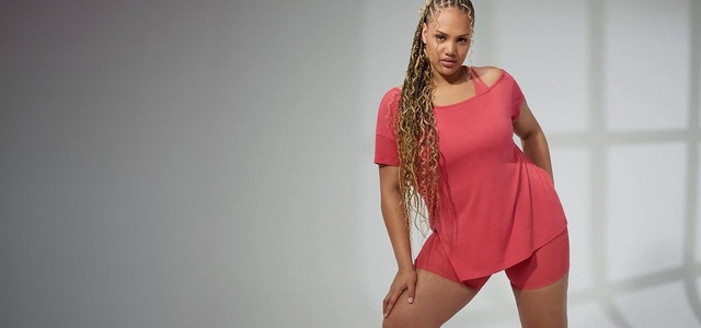A model with long braided hair poses against a light grey background wearing a coral asymmetric off-shoulder short-sleeve top with a high-low hem paired with matching coral biker shorts.