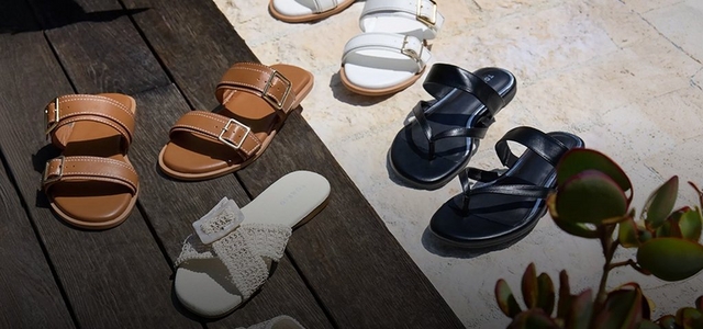 A promotional banner featuring five pairs of summer sandals arranged in a flat lay on a wooden deck and stone surface with tropical succulent foliage in the corner. Styles include tan double-strap buckle slides, white buckle slides, white crochet slides, and black strappy thong sandals.