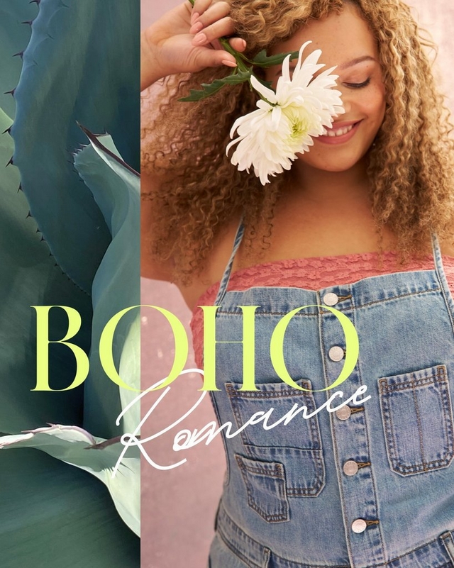 A vertical promotional banner split into two sections. The left half shows a close-up of large blue-gray agave leaves with sharp tips. The right half shows a woman with voluminous curly auburn hair smiling with eyes closed, holding a white chrysanthemum flower near her face against a dusty pink background. She wears a pink lace bandeau top under a light wash denim button-front overall dress with chest patch pockets. Overlaid across both sections, yellow-green serif text reads 'BOHO' above white script text reading 'Romance'.
