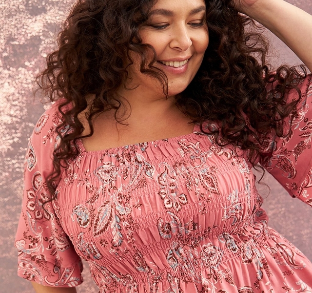 A close-up of a woman with voluminous dark curly hair smiling with her eyes cast downward, one hand raised to her head, against a textured rose-pink background. She wears a dusty rose smocked square-neck top with elbow-length flutter sleeves and a peplum hem, covered in a detailed white and gray paisley and floral print. The top features a fully smocked bodice from neckline to waist.
