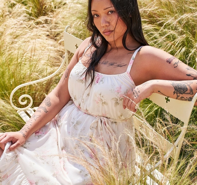 A woman with long straight dark hair sits on a white wrought iron garden chair surrounded by tall golden ornamental grass blowing in the breeze. She wears a cream spaghetti-strap maxi dress with a gathered scoop neckline, a drawstring waist tie, and a full skirt, printed with soft pink roses and green leaves. She holds the skirt out to one side. She has extensive tattoos on both arms and her chest, and looks directly at the camera with a calm expression.