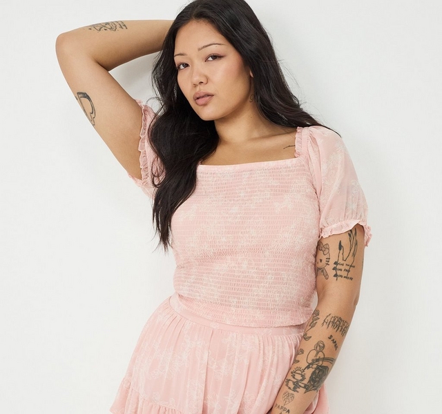 A wide horizontal banner with large green tropical leaves as the background. Two overlapping photo panels show the same woman with long straight dark hair and extensive tattoos on her arms, chest, and legs. In the left panel, she poses against a plain white background with one arm raised behind her head, wearing a light pink smocked square-neck mini dress with short puff sleeves, a fitted smocked bodice, and a tiered ruffle skirt. In the right panel, a close-up outdoor shot shows her from the chest up against a backdrop of white flowering plants and greenery, focusing on the detail of the smocked square neckline with a ruffled edge and short puff sleeves. A script chest tattoo is visible.